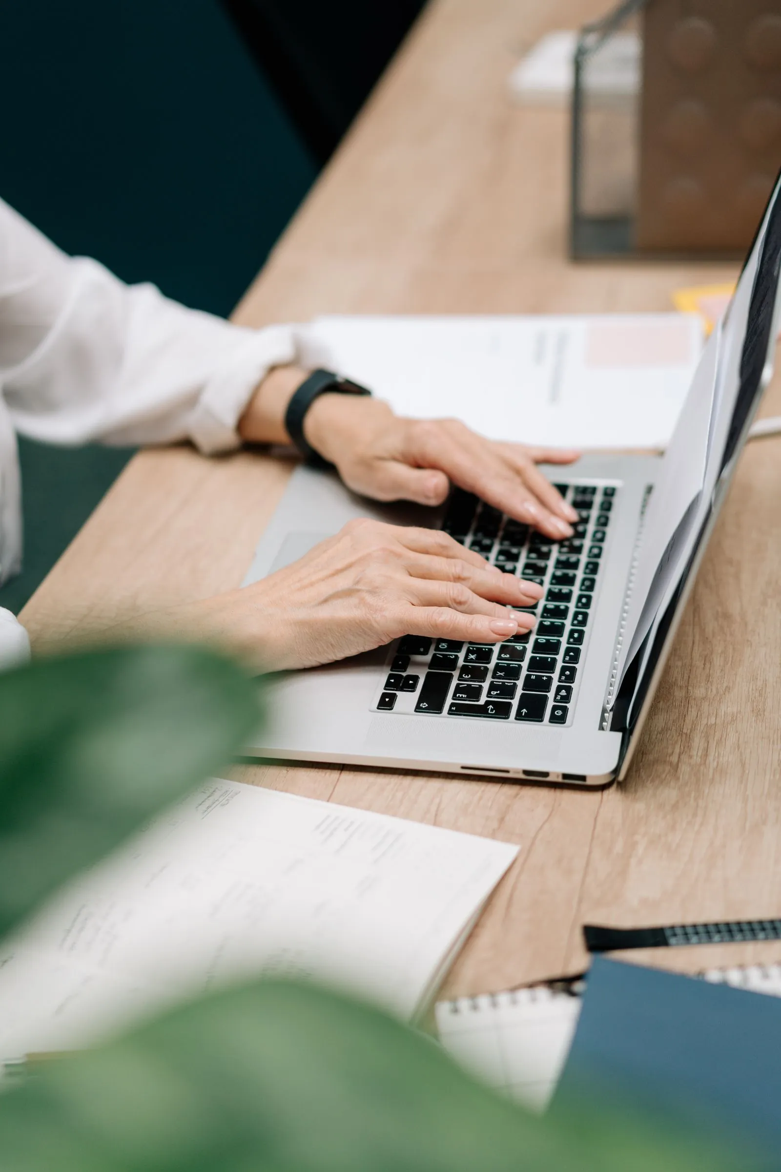Close-up of a contractor in a white shirt typing on a laptop at a wooden desk, paperwork spread beside the keyboard.