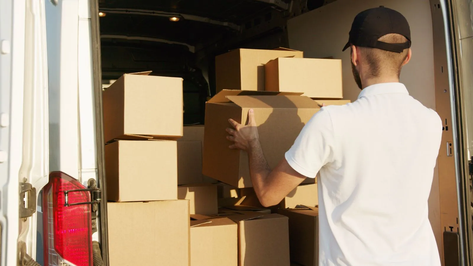 A contractor in a white polo and ball cap stacks cardboard boxes into the back of a white delivery van on a sunny morning.