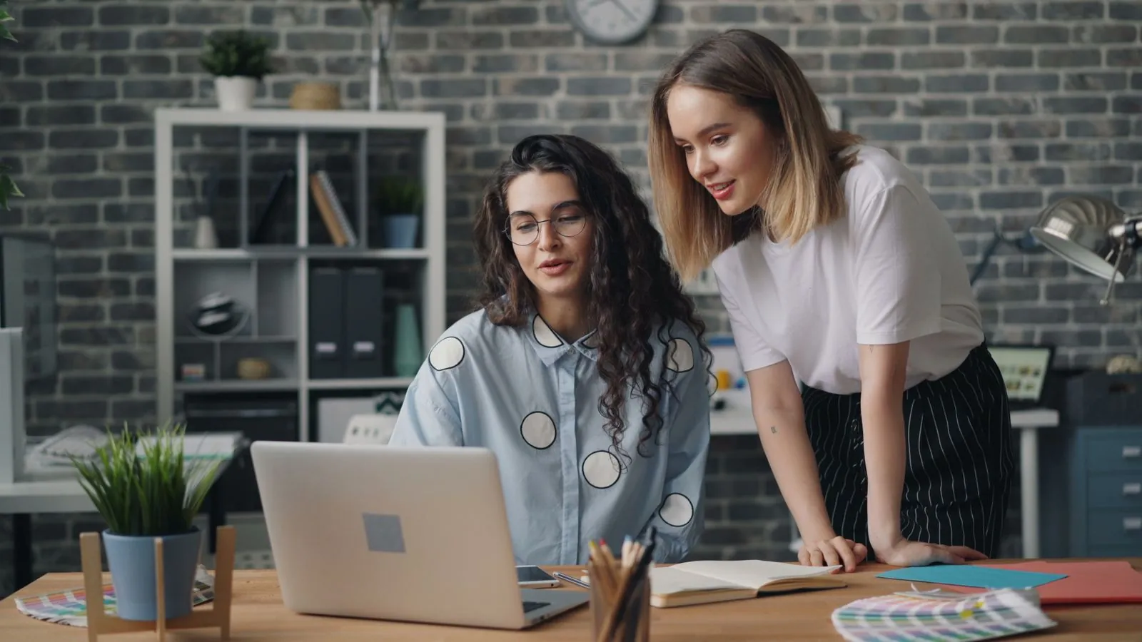 Two founders review something on a laptop together at a wooden desk in a bright brick-wall studio, mid-planning session.