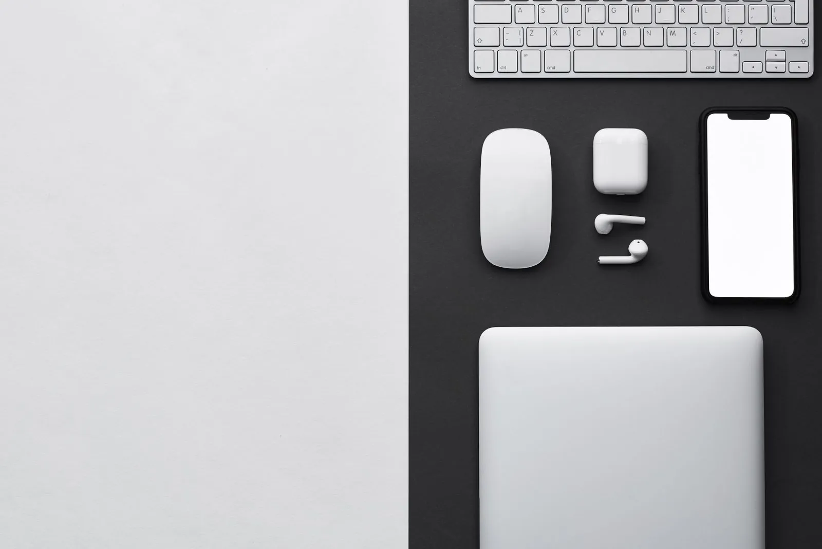 A minimalist flat lay of a laptop, keyboard, wireless mouse, earbuds, and a smartphone arranged on a two-tone black and white desktop.