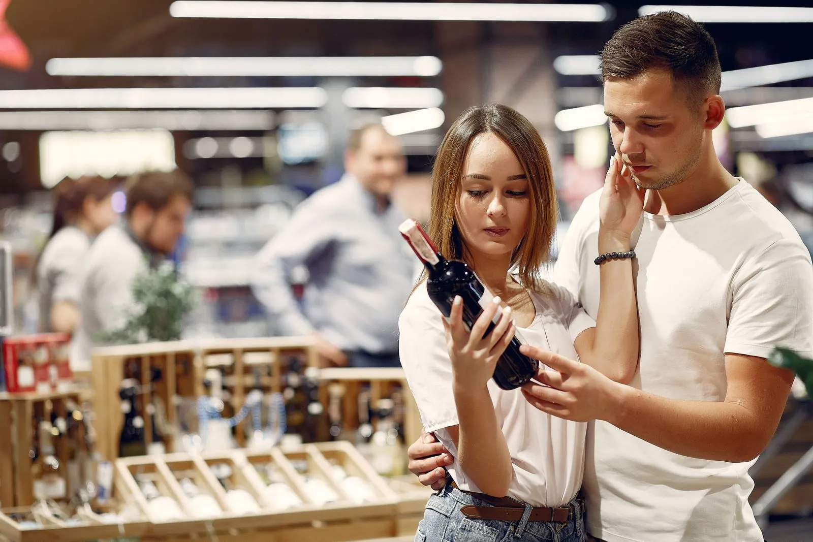 A young couple studies a wine bottle together in a grocery aisle, thoughtfully comparing labels.
