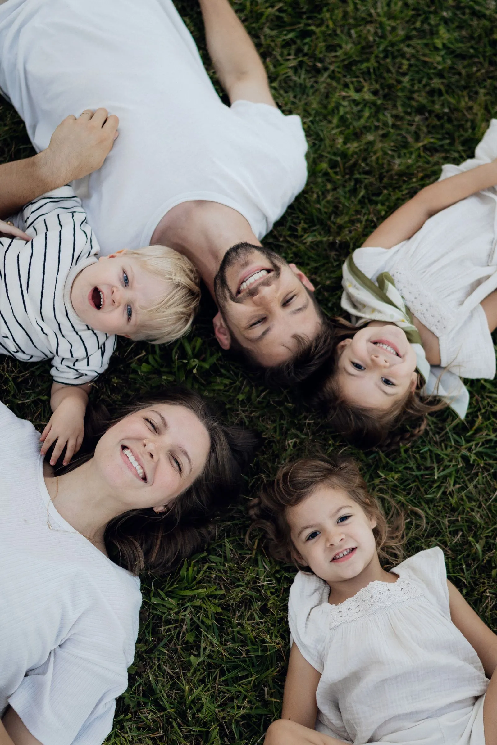 A joyful young family lies together on green grass in a circle, parents and three children smiling up at the camera.