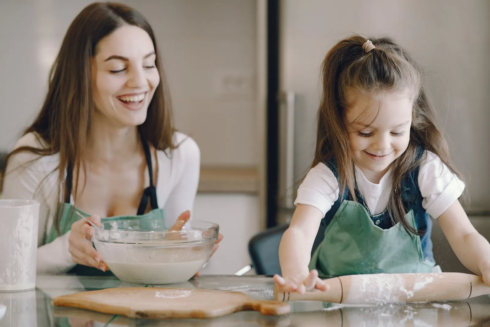 A parent and a young daughter smile together while baking in a bright kitchen, a rolling pin and bowl in front of them.
