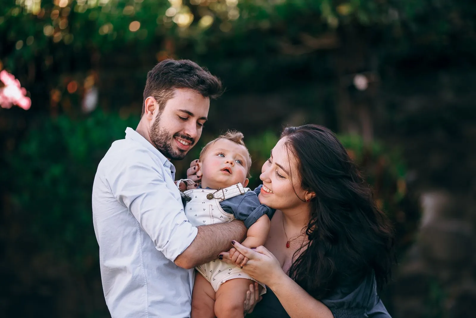 A young couple hold their smiling baby outdoors in a garden setting, warm sunset light in the trees behind them.