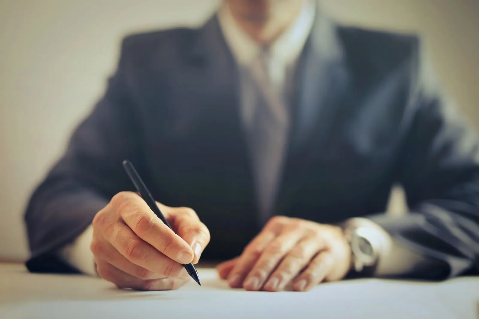 A person in a suit signs a document with a pen, the paperwork in sharp focus on the desk in front of them.