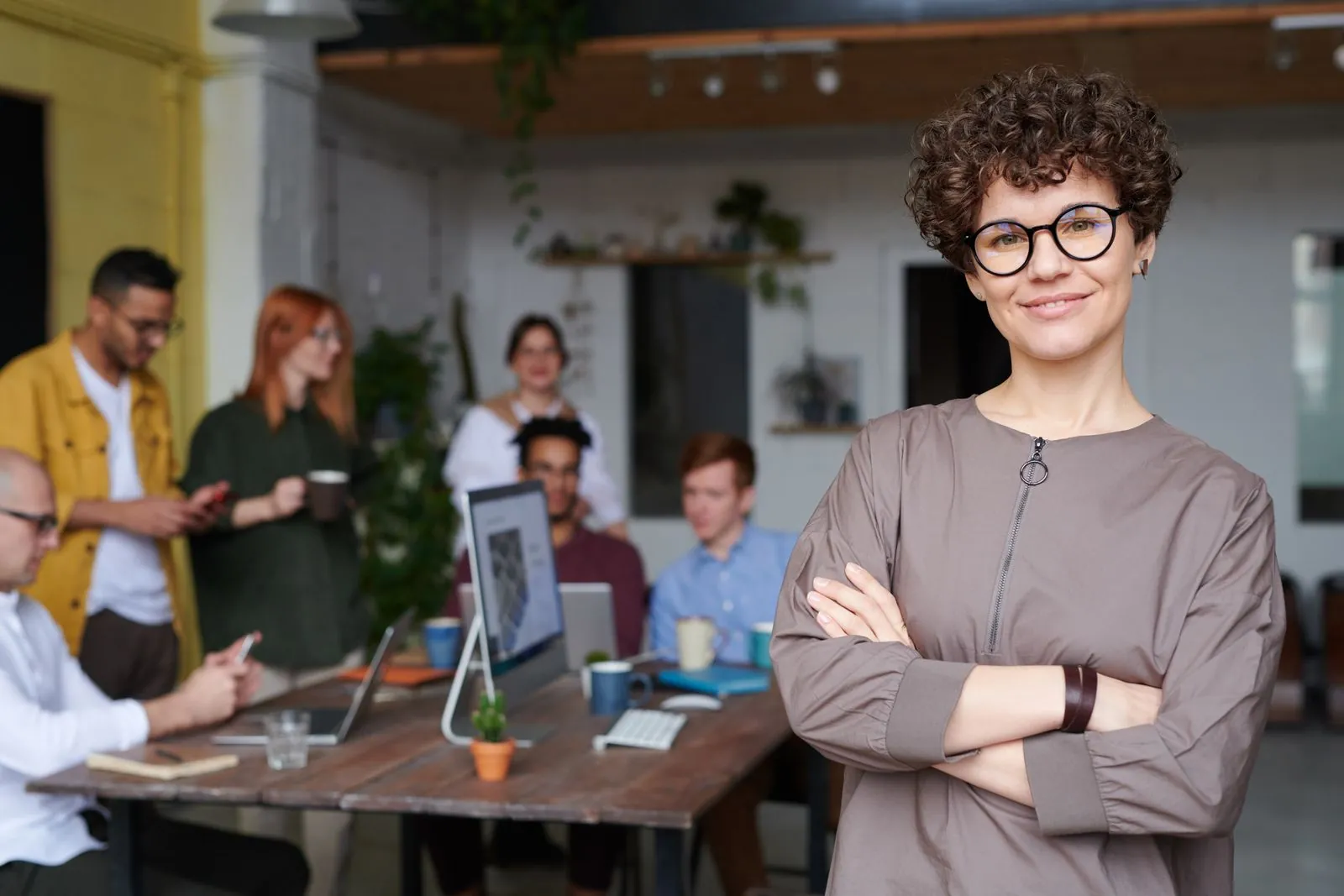 A confident professional stands with arms crossed in a modern office, smiling, colleagues collaborating on a laptop in the background.