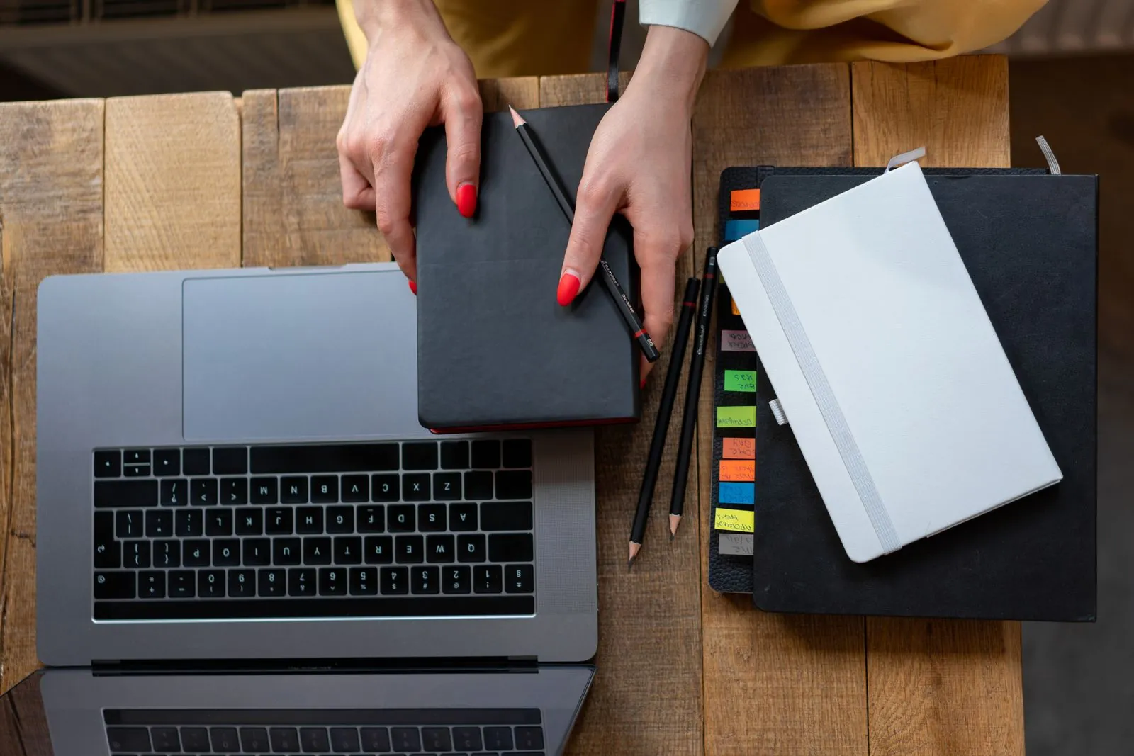 Overhead view of a person's hands organizing notebooks beside a laptop on a wooden desk in a bright workspace.