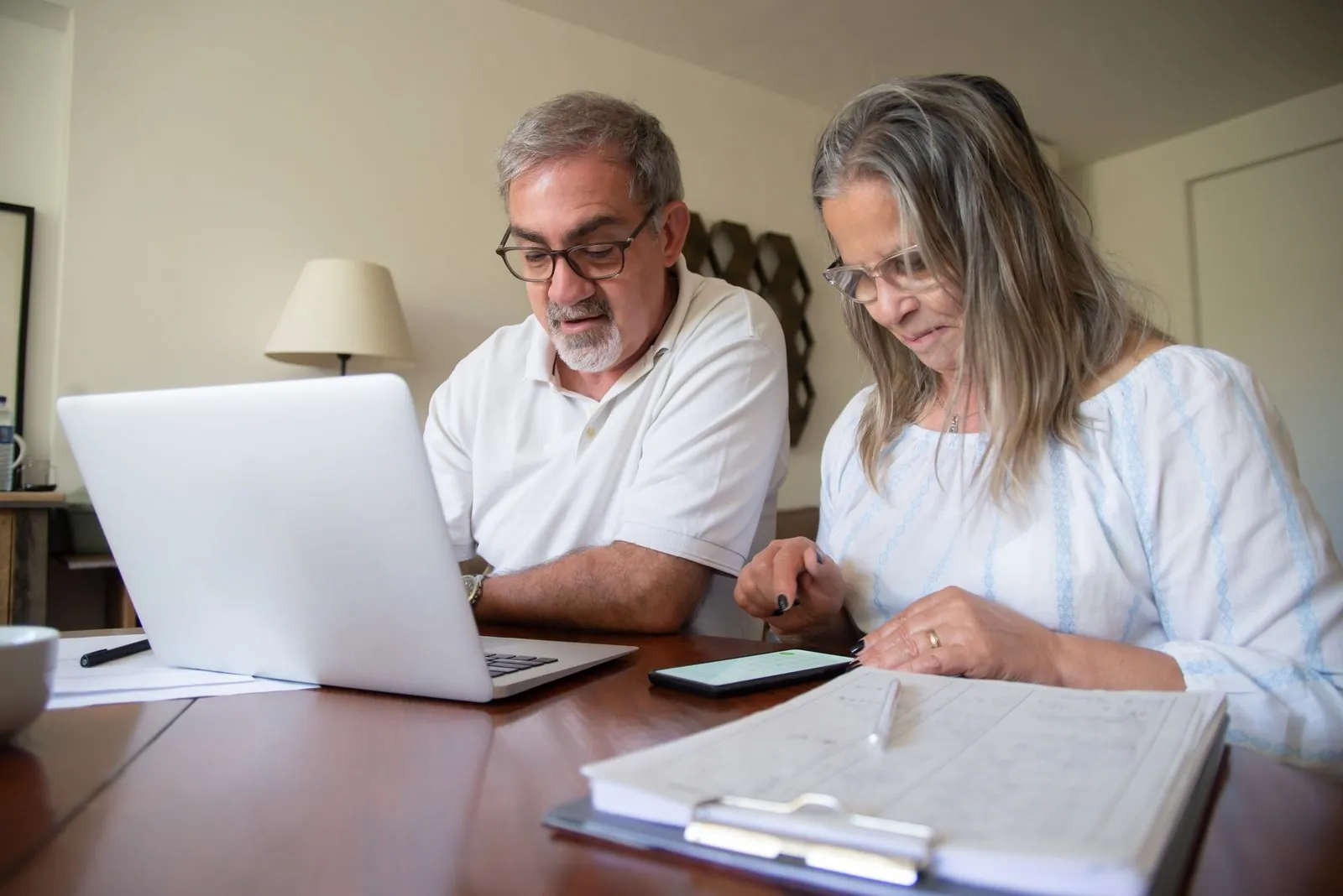 A couple sits together at a warm-lit dining table reviewing insurance paperwork with a laptop and clipboard between them.