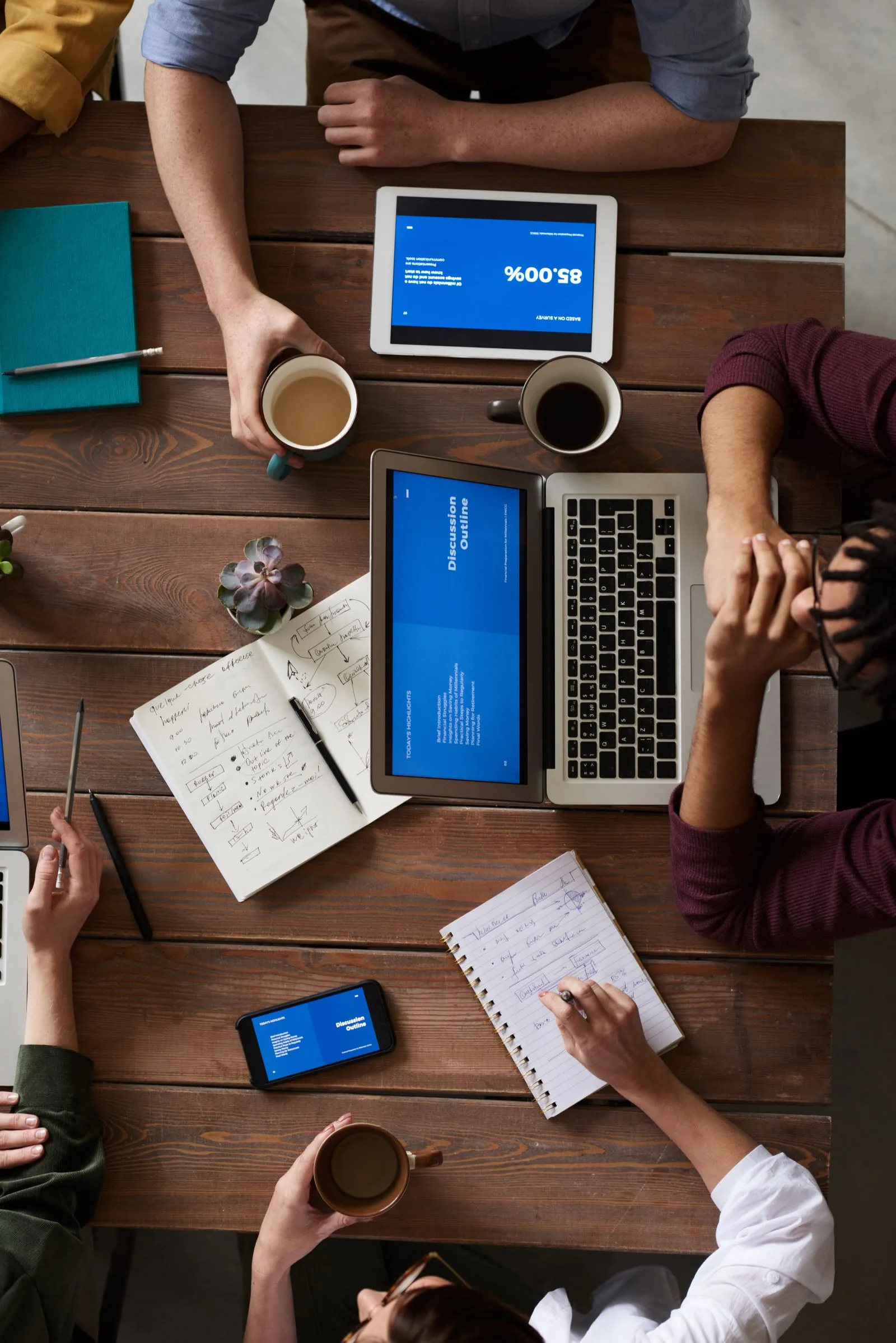 Overhead view of a team meeting around a wooden table, laptops and tablets showing charts with coffee cups and notebooks beside them.