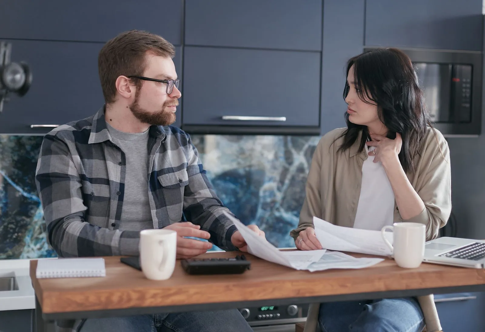 A young couple sits at their kitchen table reviewing coverage paperwork together, a laptop and mugs on the table between them.