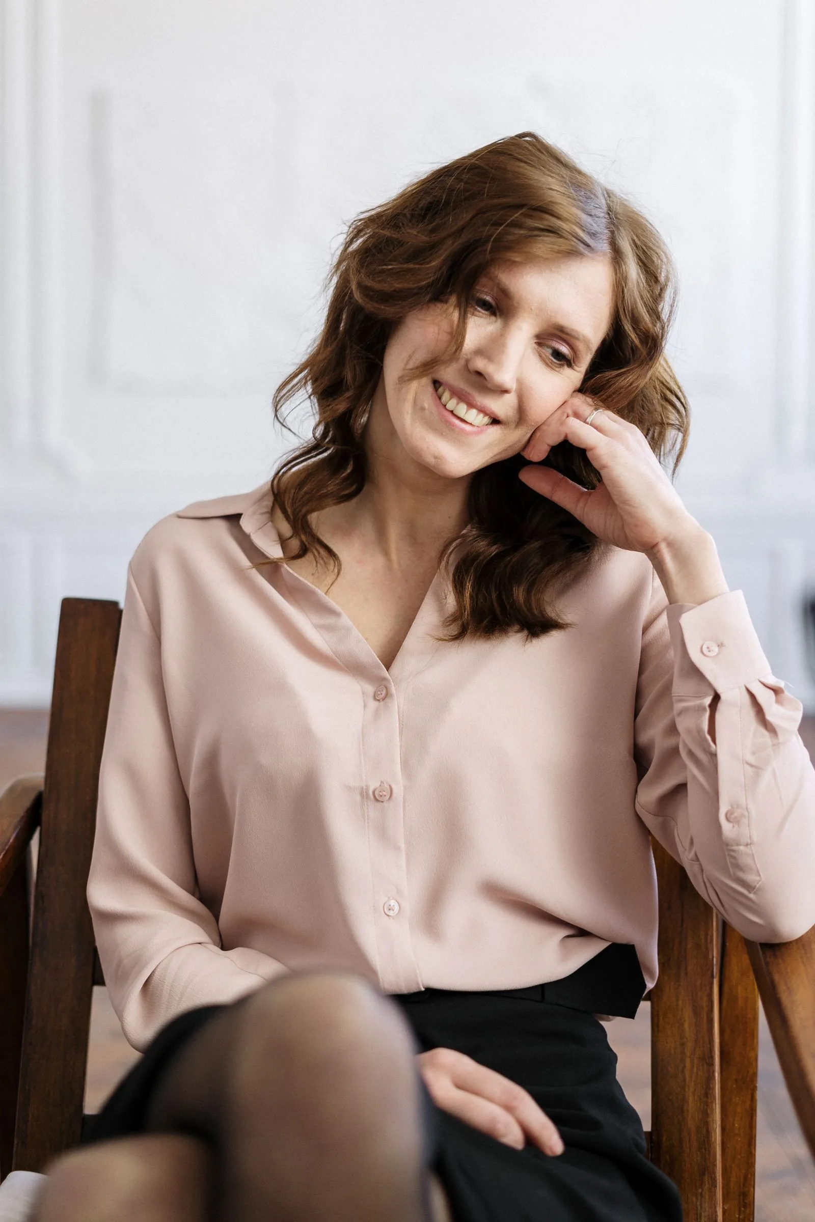 A young woman sits on a chair in a bright, minimal room, smiling gently and looking off to the side.
