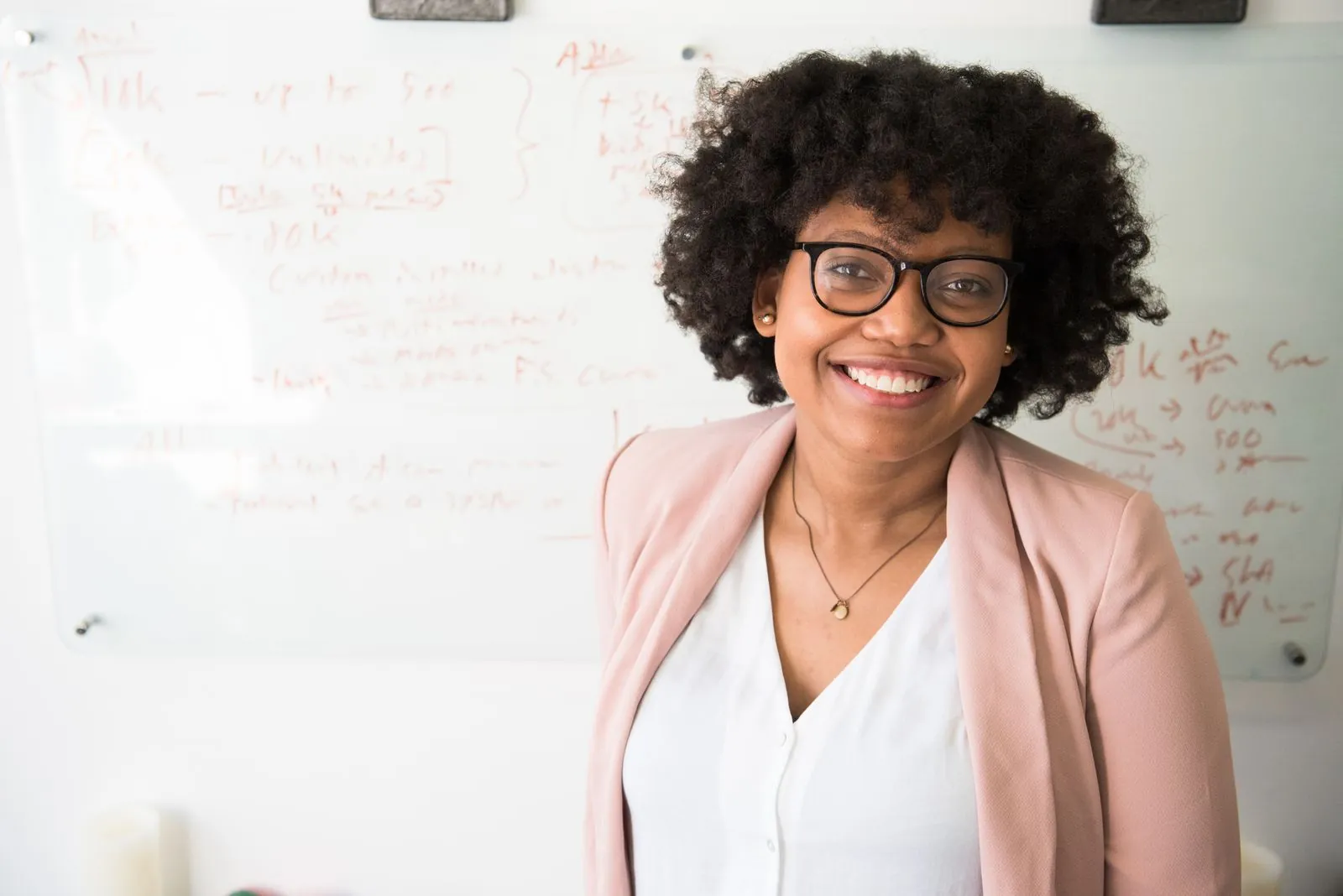 A young professional Black woman in a pink blazer smiles confidently in front of a whiteboard covered in strategy notes.