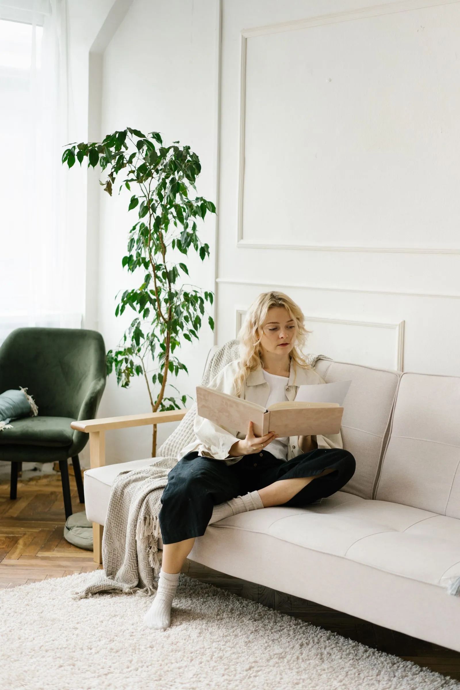 A young blonde woman sits cross-legged on a light sofa in a bright plant-filled room, quietly reading a book.