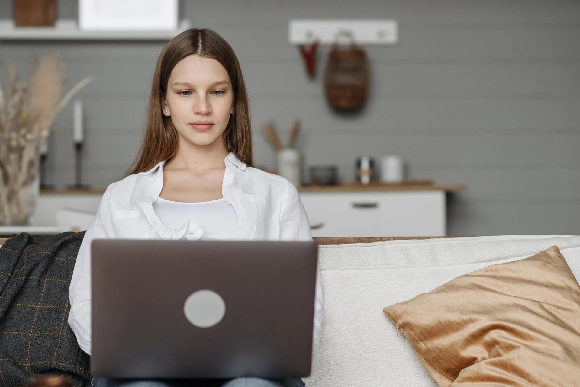 A young woman sits comfortably on a couch at home, working on her laptop with a soft, focused expression.