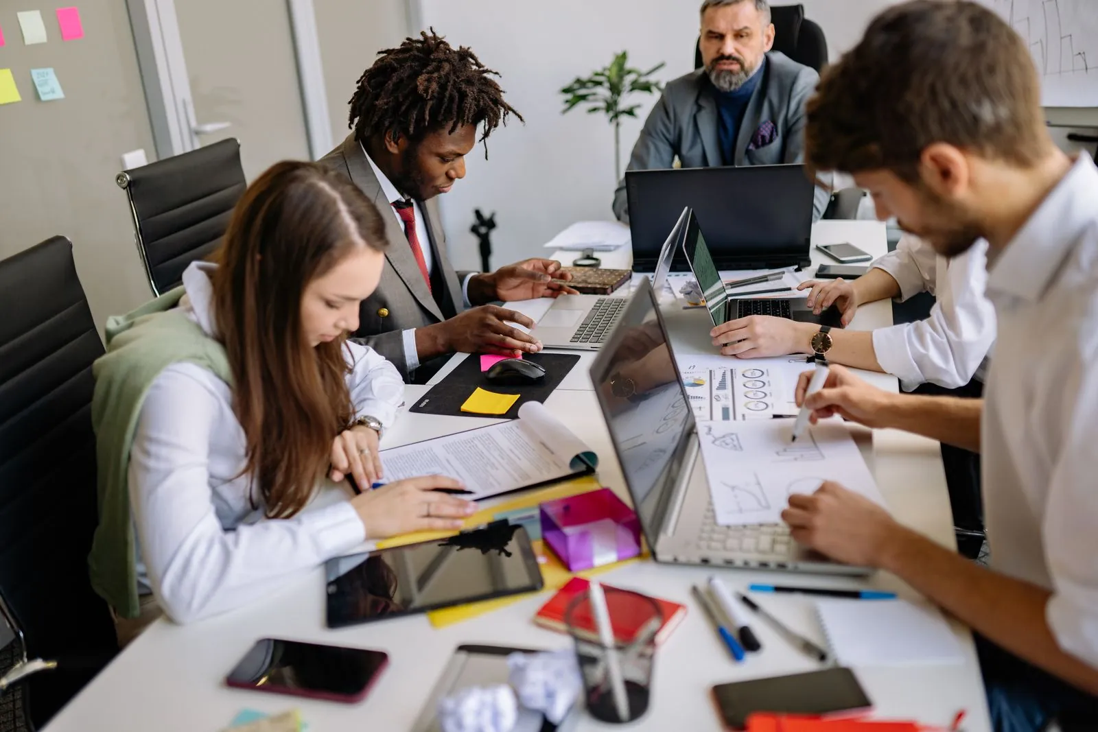 A diverse professional team collaborates around a table full of laptops and paperwork, one person taking notes and others discussing.