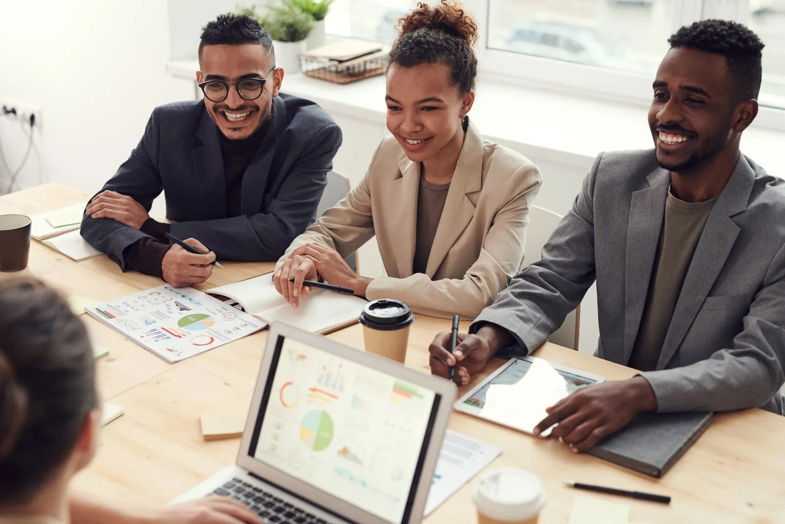 Three young professionals lean toward the camera at a bright wooden desk, smiling and holding pens, colorful charts spread out in front of them.