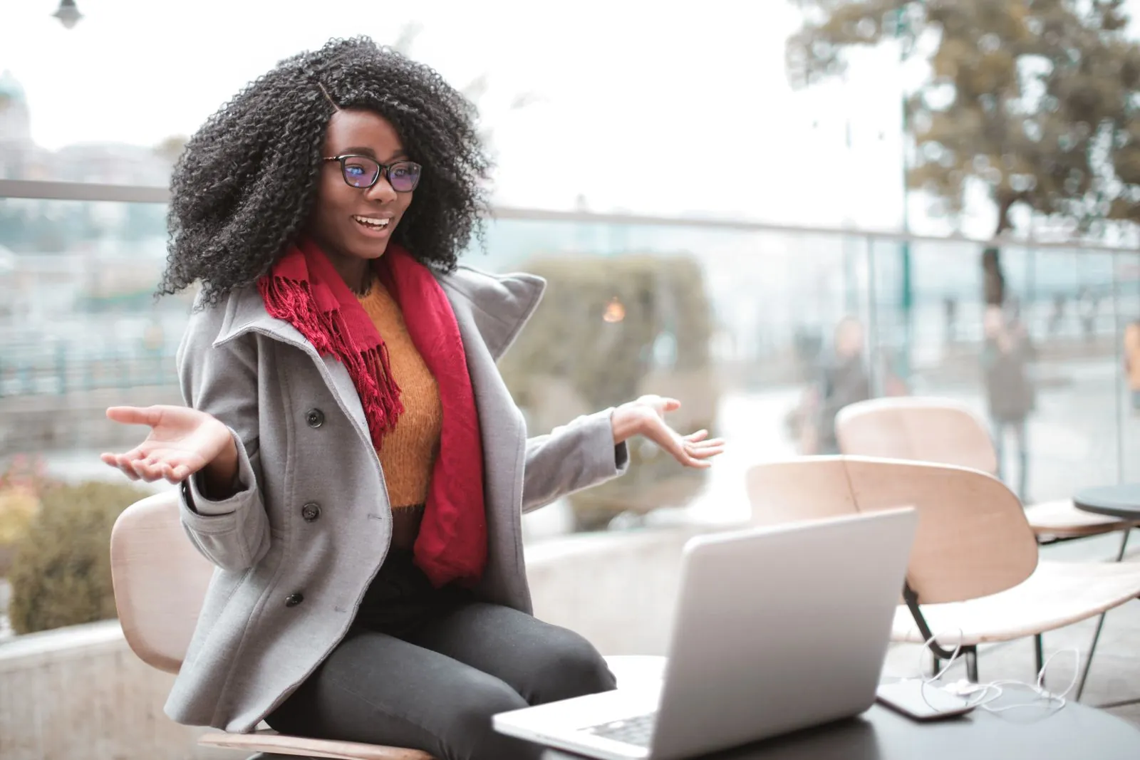 A young woman in a stylish coat sits outdoors on a balcony, laughing while gesturing at her laptop, a red scarf around her neck.