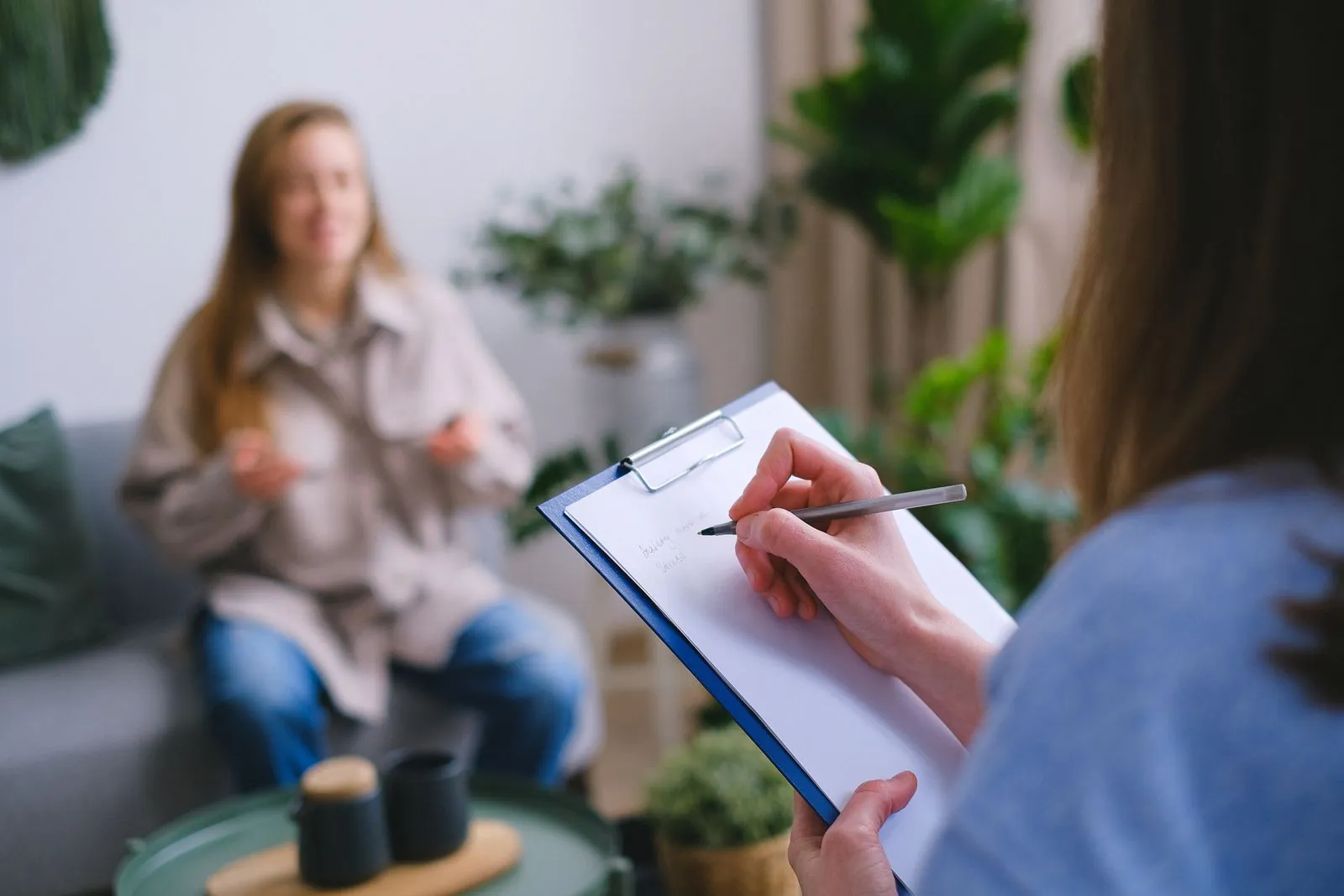 An advisor takes handwritten notes on a clipboard while listening to a client seated across the room, blurred in the background.