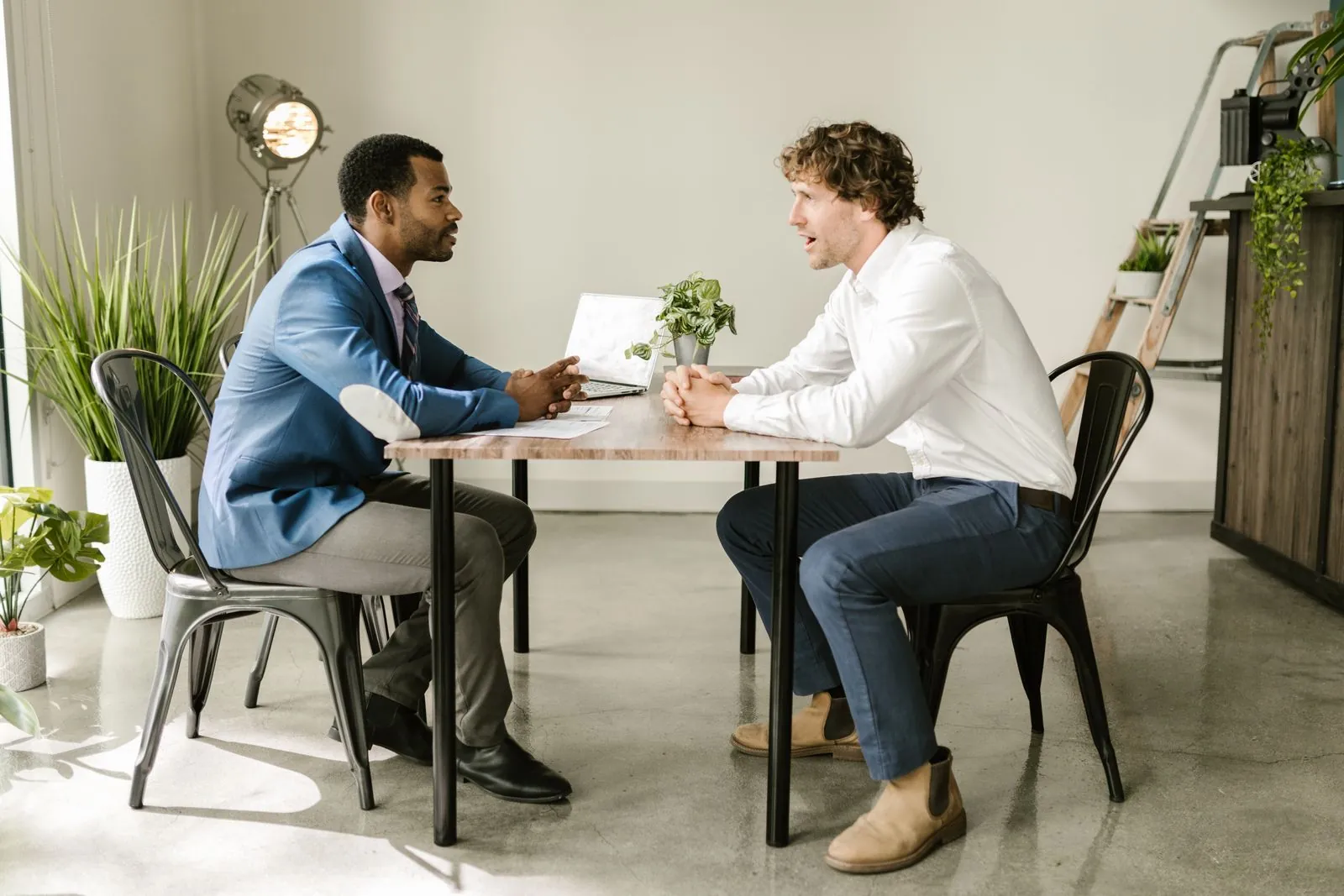 Two men sit facing each other at a small table in a bright office, one speaking with animated hands and the other listening carefully.
