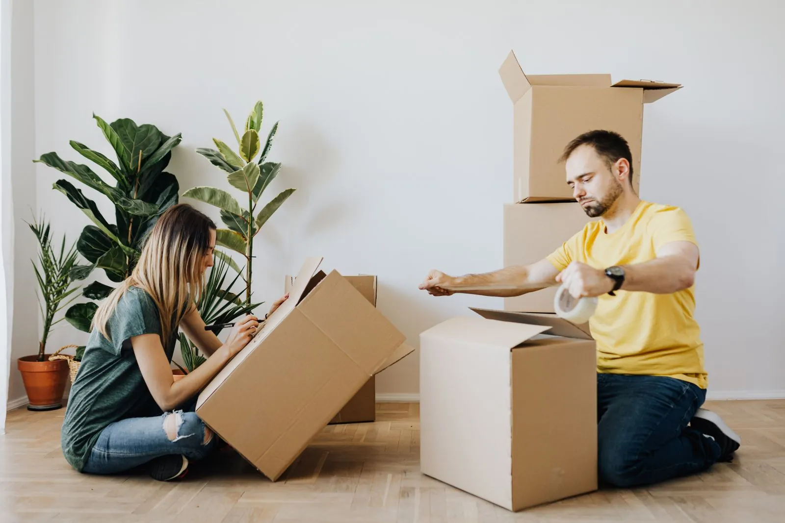A couple in a bright apartment stands surrounded by moving boxes, one carefully unpacking as the other holds tape, quietly working through the day.