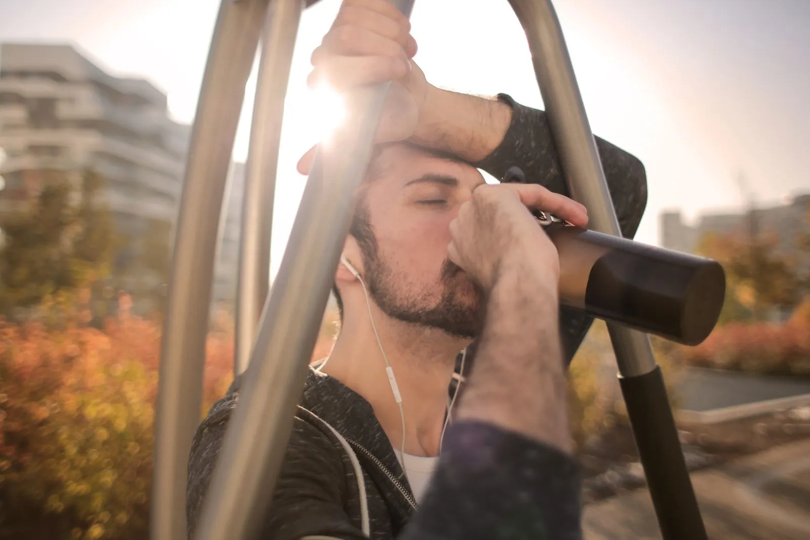 A young man exercises on outdoor gym equipment at sunset, drinking from a water bottle, city buildings behind him.